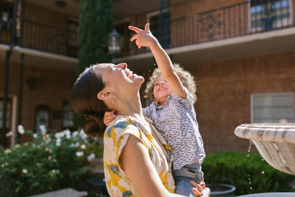 Dai “terribili 2” ai meravigliosi 3: la mia bambina che cresce troppo in fretta A mother and child share a joyful moment by a water fountain in a sunny garden setting.